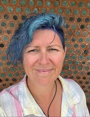 Headshot of blue haired woman with wavy curls and an undercut smiling at the camera wearing a colourful shirt and a pair of earplugs hanging around her neck for impromptu noise regulation as required!